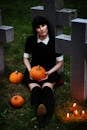 Gothic woman sitting in a graveyard holding a pumpkin surrounded by pumpkins and candles, creating a spooky Halloween vibe.